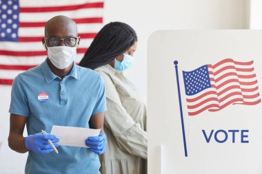Amerikan bayrağıyla süslenmiş ve salgın sonrası seçim gününde kameraya bakan Afro-Amerikalı bir adamın belden yukarı portresi.