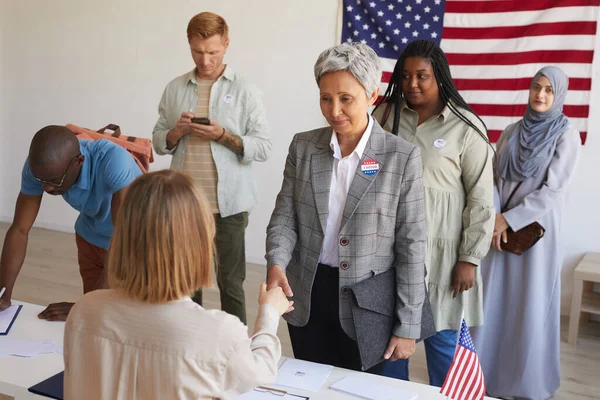 投票所の多民族グループは 選挙の日にアメリカの国旗で飾られ 投票関係者と握手する高齢女性の笑顔に焦点を当て スペースをコピーします — ストック写真