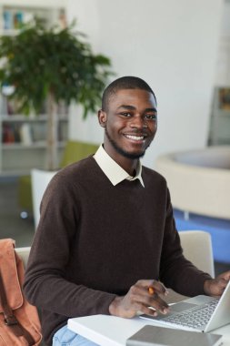 Üniversite kütüphanesinde arka planda insanlarla birlikte dizüstü bilgisayar kullanırken kameraya gülümseyen genç Afro-Amerikan bir adamın dikey portresi