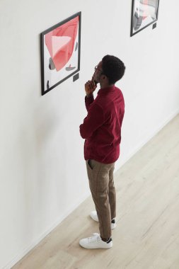 Tablolara bakan ve sanat galerisi ya da müze sergisinde düşünen Afro-Amerikan genç bir adamın dikey yüksek açılı portresi.
