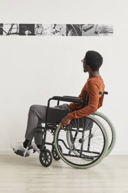 Tekerlekli sandalye kullanan ve modern sanat galerisi sergisini keşfederken resimlere bakan genç bir Afro-Amerikalının tam boy portresi.
