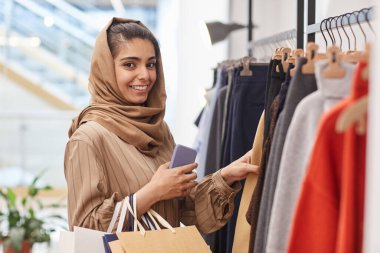 Elbise askılarının yanında durup alışveriş merkezinden alışveriş yaparken kameraya bakan zarif genç bir kadının belini kaldırın.