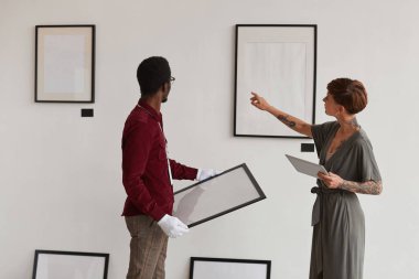 Kadın sanat galerisi müdürünün arka plan portresi müzede sergi planlarken işçilere beyaz duvarlara çerçeve asma talimatı veriyor.