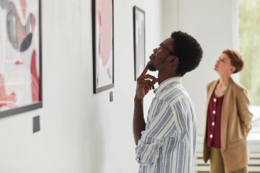 Modern sanat galerisi sergisini keşfederken resimlere bakan Afro-Amerikan genç bir adamın yan görünüm portresi.