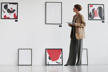 Modern sanat galerisini ziyaret ederken beyaz duvara asılı resimlere bakan zarif bir kadının tam boy portresi.