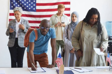 Seçim günü Amerikan bayraklarıyla süslenmiş, oy pusulasına kayıtlı çok ırklı bir grup insan oy pusulası imzalayan Afrikalı adama odaklanıyor ve kameraya bakıyor, uzay boşluğunu kopyalıyor.