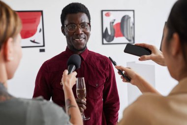 Modern sanat galerisi açılışı sırasında gazetecilere röportaj veren gülümseyen Afro-Amerikan bir adamın belden yukarı portresi.