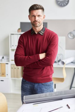 Yetişkin sakallı bir mimarın dikey portresi. Kolları çapraz duruyor ve iş yerinde masa çizerken kameraya bakıyor.