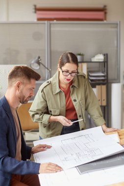 İki mimarın dikey portresi planlara bakıyor ve modern ofiste masanın başında dururken iş tartışıyorlar.