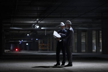 İnşaat alanında karanlıkta dikilirken, şapka takan ve planları tutan iki iş adamının dramatik uzun portresi.