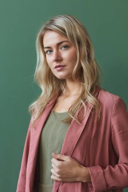 Tozlu pembe ceket giyen, stüdyoda yeşil arka planda poz verirken kameraya bakan zarif sarışın kadının dikey bel portresi.