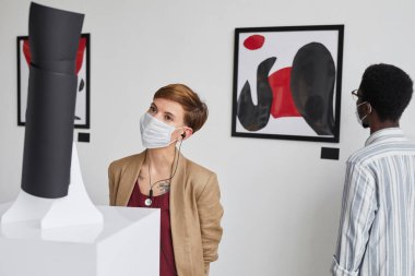 Maske takıp modern sanat galerisini keşfederken resim ve heykellere bakan iki gencin portresi.