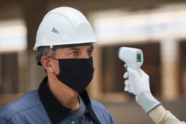 Maske takan olgun bir işçinin portresi ve inşaat sahasındaki temassız termometreyle sıcaklığı ölçmek için bekliyor. Corona virüsü güvenliği.