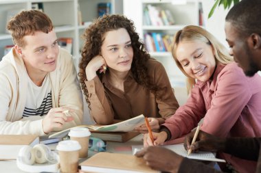 Üniversite kütüphanesinde masada oturup grup projesi üzerinde çalışırken birlikte çalışan çok ırklı, gülümseyen gençler grubu.