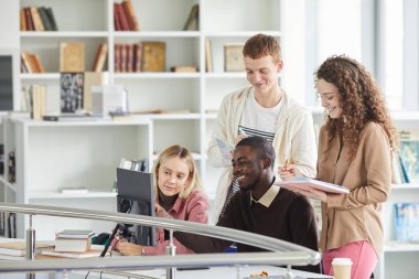 Kolej kütüphanesinde okurken telekomünikasyon ekipmanı kullanan ve gülümseyen çok ırklı bir öğrenci grubu.