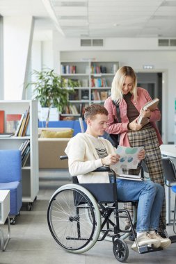 Kolej kütüphanesinde genç bir kadınla konuşan neşeli engelli öğrencinin dikey portresi