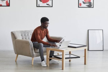 Modern sanatla dekore edilmiş kanepede otururken dizüstü bilgisayarda çalışan genç Afro-Amerikan bir adamın portresi.