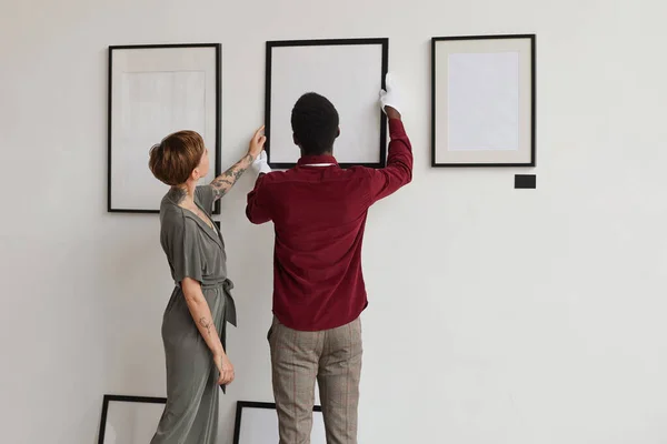 stock image Back view portrait of two art gallery workers hanging painting frames on white wall while planning exhibition in museum, copy space