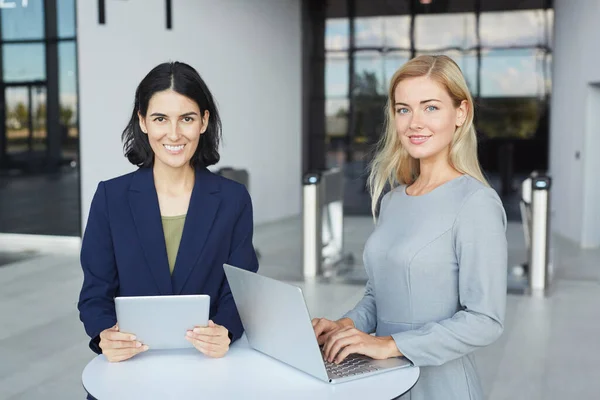 2名成功的女商人站在办公大楼的办公桌前 手持笔记本电脑 一边举着相机 一边举着画像 一边对着镜头笑 — 图库照片