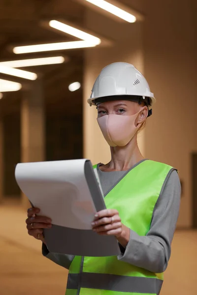 Vertical waist up portrait of mature female worker wearing mask and looking at camera while standing with clipboard at construction site indoors