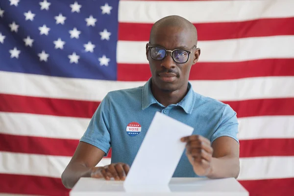 投票箱に投票用紙を入れて 選挙の日にアメリカの旗に対して立っている間にカメラを見て 現代のアフリカ系アメリカ人の男の肖像画 スペースをコピー — ストック写真