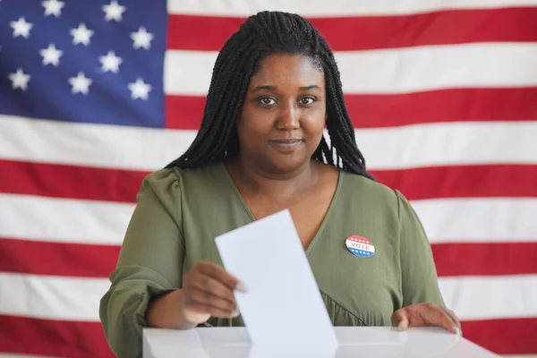 投票箱の中に投票用紙を置き 選挙の日にアメリカの国旗に反対しながらカメラを見る若いアフリカ系アメリカ人女性の肖像画 スペースをコピー — ストック写真