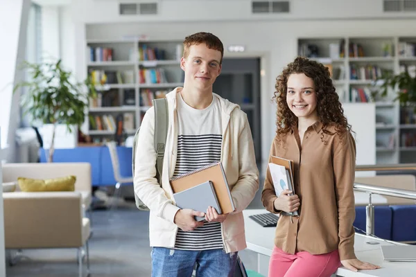 Waist up portrait of young student couple looking at camera and smiling while standing in college library holding books, copy space