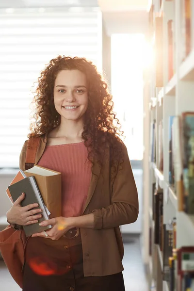 Vertical waist up portrait of young woman holding books and smiling at camera while standing in school library