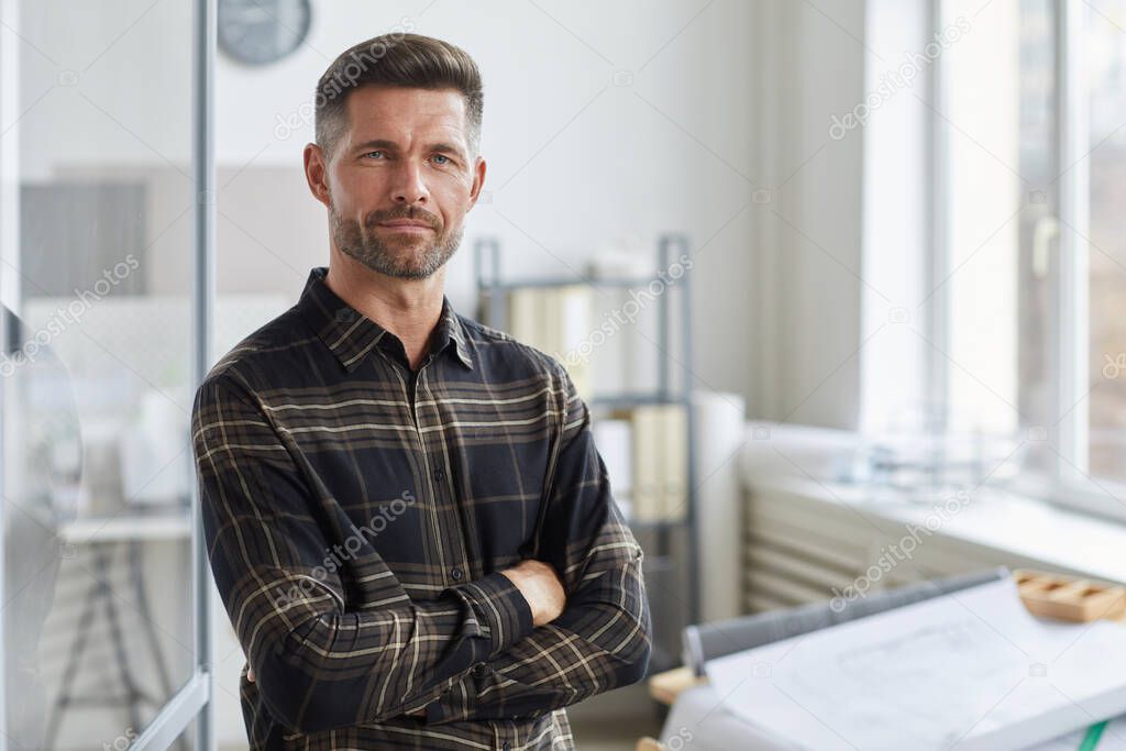 Waist up portrait of confident bearded architect looking at camera while standing with arms crossed by drawing desk in office, copy space