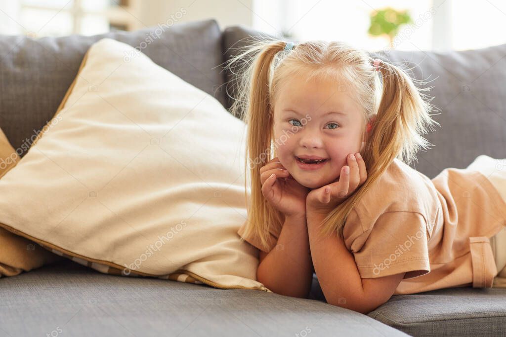 Portrait of cute girl with down syndrome smiling happily at camera while lying on sofa at home, copy space