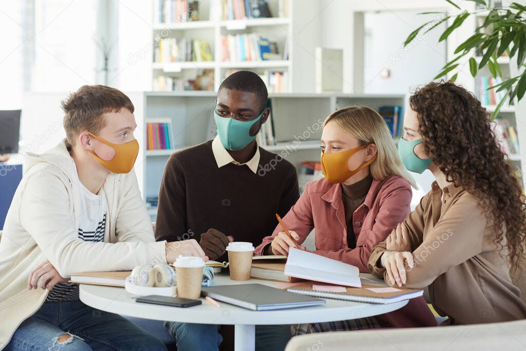 Multi-ethnic group of young people wearing masks while studying together at table in college library, copy space
