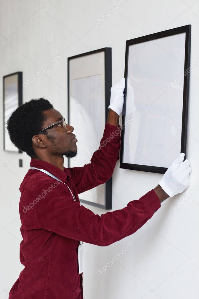 Vertical side view at African-American man hanging frames on wall while planning art gallery or exhibition