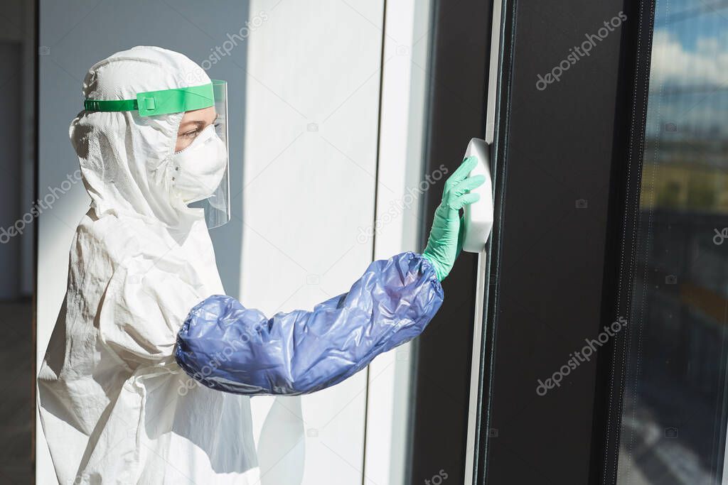 Side view portrait of female worker wearing hazmat suit disinfecting windows in office building, copy space