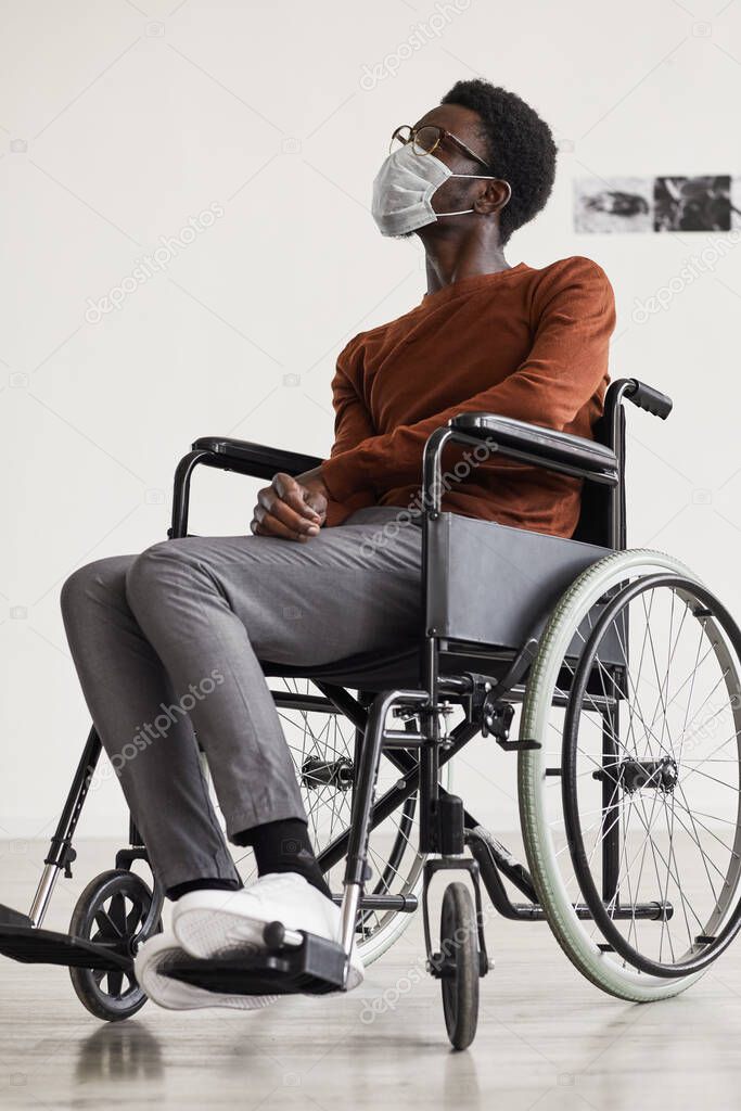 Vertical full length portrait of young African-American man using wheelchair and wearing mask while looking at paintings in modern art gallery