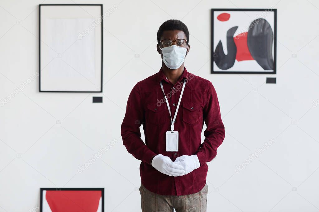 Front view portrait of African-American man wearing mask looking at camera while standing against modern graphic paintings in art gallery, copy space