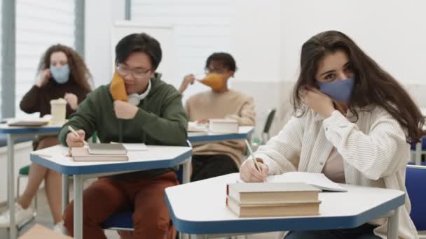 不同的学生坐在课桌旁 在笔记本上写字 摘下五颜六色的口罩 举起口罩 混血儿望着 对着镜头笑 — 图库视频影像