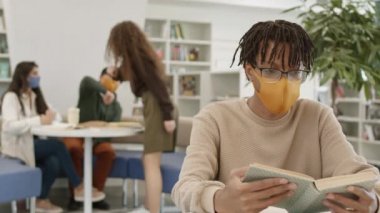 Sarı bez maskeli zeki Afrikalı adamın kütüphanede kitap okuyup sonra da kameraya baktığı yakın çekim görüntüsü. Bulanık öğrenciler öğreniyor, arka plandaki koltukta oturuyorlar.