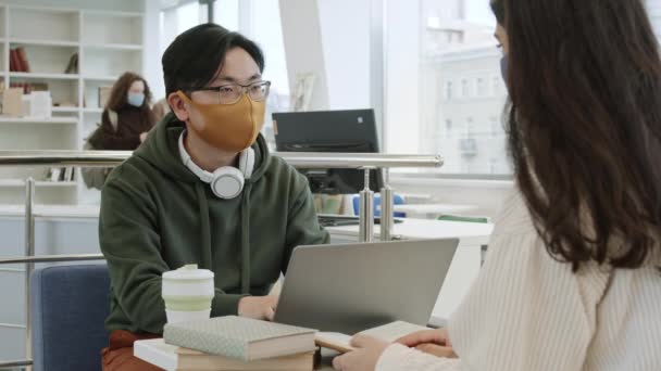 在明亮的图书馆里 戴着面具和眼镜坐在笔记本电脑上打字的聪明的亚洲男人肩膀上的照片 与难以辨认的女性朋友 走在后面的人交谈 一起做项目的学生 — 图库视频影像
