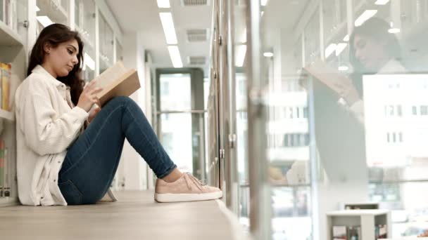混血女学生坐在地板上 靠着书架 在公共图书馆看书 然后转过头 对着镜头微笑 全景全景全景拍摄 — 图库视频影像