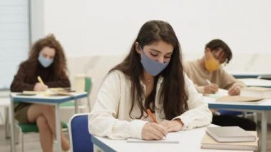 Karışık ırk üniversiteli bir kızın göğsü açılır. Masa başında oturup derste deftere yazı yazar. Öğrenciler salgın sırasında maske takarak öğreniyor.