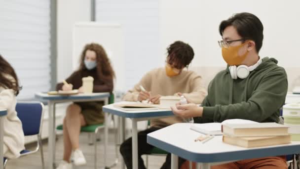 Ázsiai Fickó Szemüvegben Kendőmaszkban Asztalnál Osztályteremben Fertőtlenítő Folyadékot Permetez Kezére — Stock videók