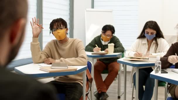 フェイスマスクをした多様な学生たちが 教室の机の上で講義をしている姿を肩から撮影 アフリカ人の男が手を挙げて正体不明の教授に質問する — ストック動画