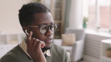 Genç Afro-Amerikalı iş adamının gözlük taktığı ve kablosuz kulaklık taktığı orta boy bir fotoğraf. İş yerinde oturuyor ve kulaklıkla telefon alıyor.