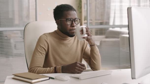 若いアフリカ系アメリカ人男性のオフィスの労働者の中程度のショットオフィスや飲料のデスクトップに座ってカジュアルな服や眼鏡を着用 — ストック動画