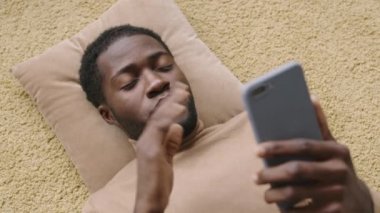 Genç Afro-Amerikalı iş adamının şık giysiler giyip, halının üzerinde yatıp, telefon kullanarak video konuşması yaptığı yüksek açı.