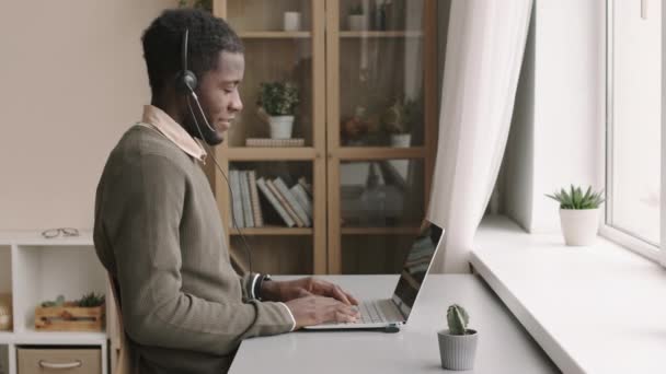 若いアフリカ系アメリカ人男性コールセンター労働者の中ショットノートパソコンの前にデスクトップに座ってビデオ通話に答えるヘッドセットを身に着けている — ストック動画