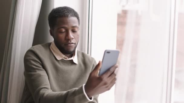 Lockdown Young African Man Wearing Casual Clothes Sitting Windowsill Mobile — Stock Video