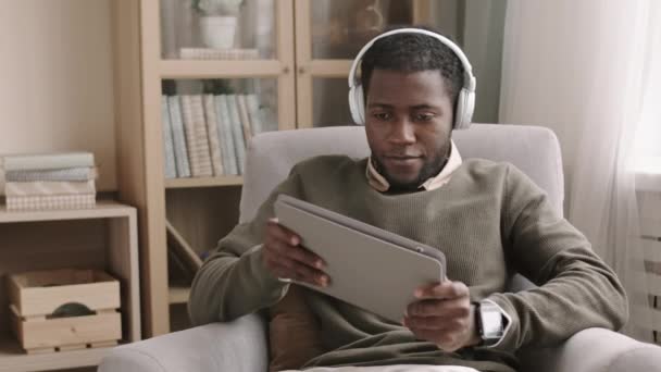 彼の手にタブレットを保持し ビデオゲームをプレイし 椅子に座ってカジュアルな服やヘッドフォンを身に着けている若いアフリカ系アメリカ人の男の中のショット — ストック動画