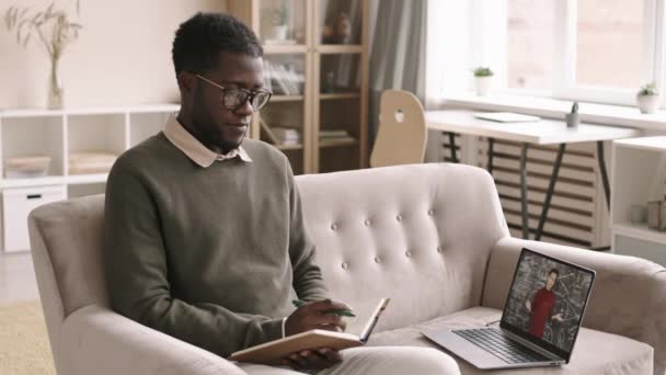 若いアフリカ系アメリカ人の男のロックダウンカジュアル服や眼鏡を自宅でソファに座って ノートパソコンを使用してビデオ天文学のレッスンを受けながら眠りに落ちる — ストック動画