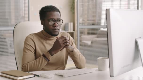 オフィスの職場に座ってコンピュータの画面を見て同僚と遠くに話をするカジュアルな服や眼鏡を身に着けている若いアフリカの男性のオフィスの労働者の中程度のショット — ストック動画